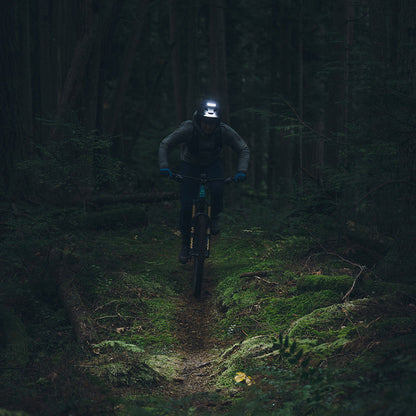 Mountain biker riding a dark forest trail lit by Outbound bike lights, including the Portal helmet light